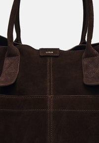 Sac cabas en daim marron foncé avec deux poignées courtes, coutures contrastantes et une étiquette de marque visible portant l'inscription "SOEUR". Design minimaliste.