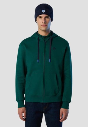 Uomo che indossa una felpa con cappuccio verde scuro con cerniera e cordini neri, e un berretto blu navy, in piedi contro uno sfondo chiaro e uniforme.