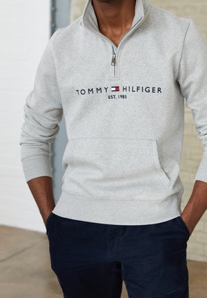 Mężczyzna w jasnoszarej bluzie Tommy Hilfiger z zamkiem do połowy i logo oraz w granatowych spodniach, z rękami w kieszeniach.