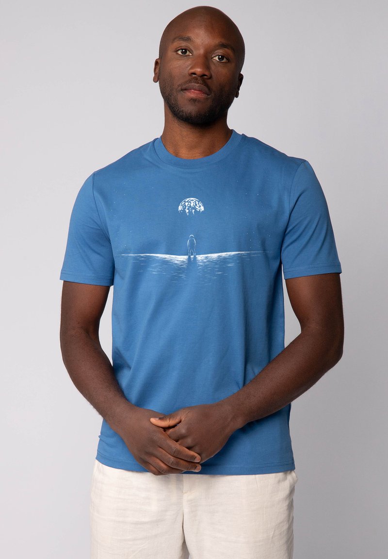 Blauwe katoenen t-shirt met een grafisch ontwerp van een persoon die bij water staat onder een gestileerde lucht met abstracte elementen. Korte mouwen.