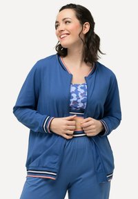 Giacca sportiva blu con polsini e colletto a righe, tessuto leggero, vestibilità comoda. Sotto, è visibile un crop top blu con motivo.
