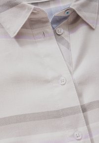 Gros plan d'une chemise beige à col avec des rayures horizontales subtiles et trois boutons blancs le long de la patte de boutonnage.