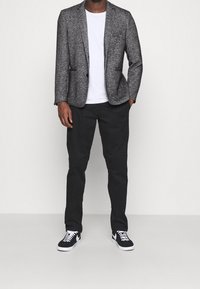 Grijze gestructureerde blazer over een wit T-shirt, gecombineerd met zwarte broek en zwarte sneakers met witte accenten, staand tegen een neutrale achtergrond.