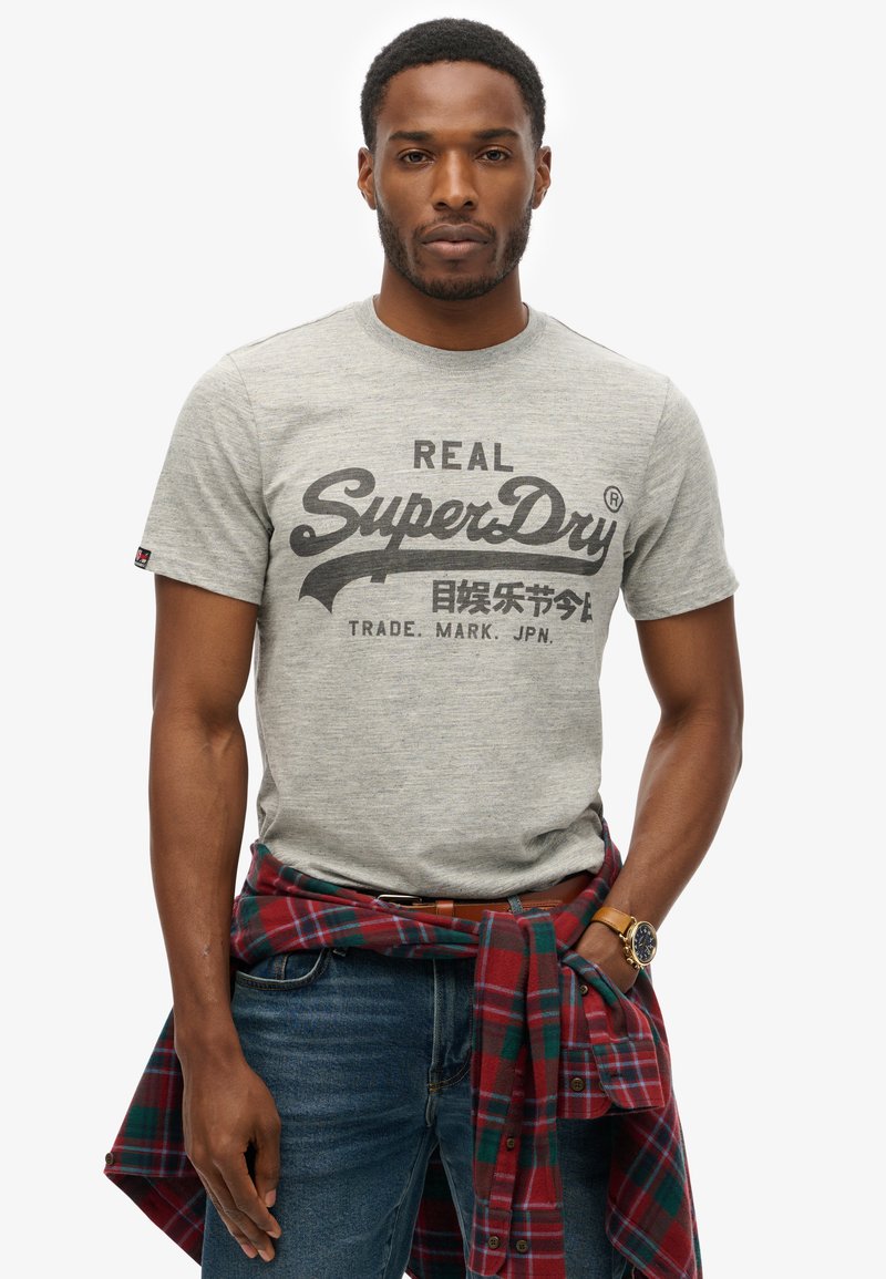Camiseta de algodón gris con el estampado negro "SuperDry". Se lleva con jeans azules, una camisa de cuadros roja y verde atada a la cintura, y un reloj marrón.
