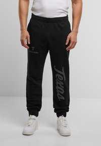 Sort sweatpants med elastisk talje, med trykt tekst "Texas" og "Upscale Studios" i kontrastfarve grå. Båret sammen med hvide sneakers.
