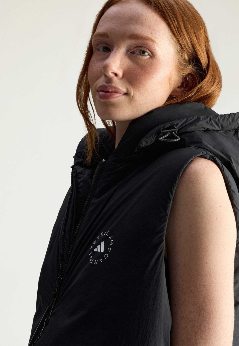 Zwarte mouwloze puffer vest met een hoge kraag, voorzien van een wit logo op de borst en een gladde, glanzende textuur. Ritsdetails zichtbaar.