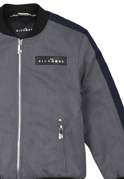 Giacca bomber grigia con accenti neri. Presenta una zip frontale, tasche laterali e una toppa del logo sul petto. Texture liscia.