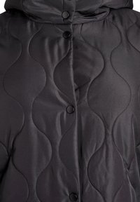 Melnas polsterētas pūkaina jaka ar vilnveida raksta šuvumiem, gludu faktūru un melnām spiedpogām priekšējā aizdares vietā.