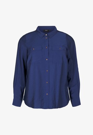 Námorně modrá košile na knoflíky s texturovaným povrchem, dvěma kapsami na hrudi a hnědými knoflíky. Má klasický límec a dlouhé rukávy.