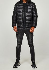 Zwarte gewatteerde jas met glanzende afwerking, horizontale doorgestikte lijnen en ritssluiting. Gecombineerd met versleten zwarte skinny jeans en sportieve sneakers.