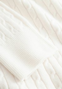 Detaliu al unui pulover cu torsade de culoare bej deschis, prezentând un model texturat cu dungi ridicate. Material moale, cu un finisaj neted și margini rotunjite.