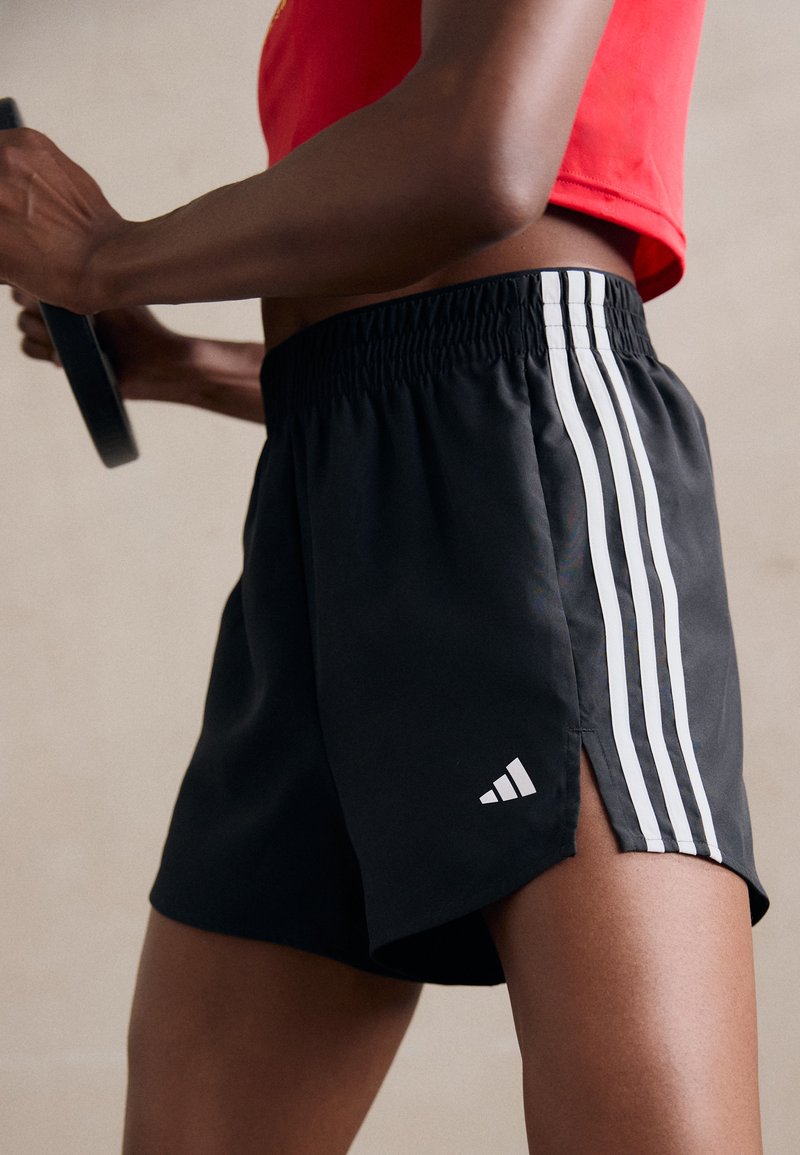 Black athletic shorts with a side slit, featuring three white stripes on each side and a small white logo on the hem. Lightweight fabric.