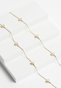 Goldene Kettenarmband mit abwechselnden herzförmigen Gliedern, mit einer glatten, polierten Oberfläche, auf einer schlichten weißen Fläche liegend.