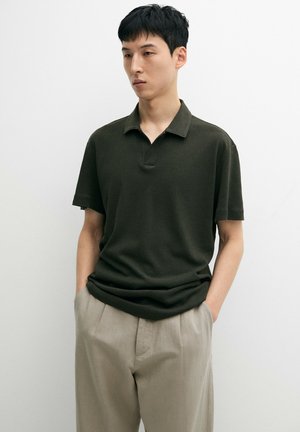 Junger Mann trägt ein dunkelgrünes kurzärmliges Poloshirt und hellbeige Hose, mit den Händen in den Taschen stehend vor einem einfarbigen weißen Hintergrund.