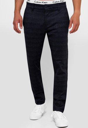 Mann trägt schwarz-graue karierte Hose, sichtbarer weißer Calvin Klein Bund, kombiniert mit weißen Sneakers, steht vor einem einfarbigen Hintergrund.