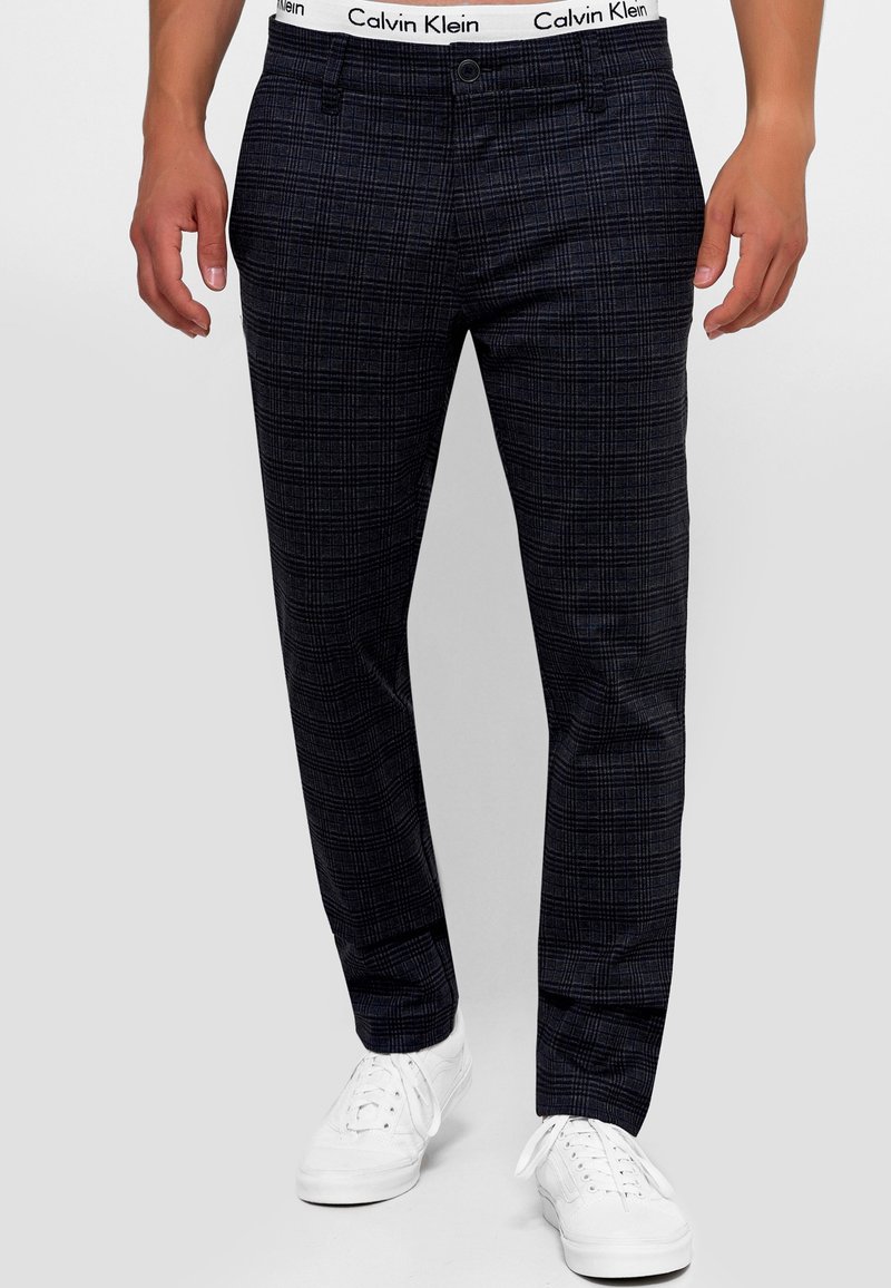 Homme portant un pantalon à carreaux noir et gris, taille blanche Calvin Klein visible, assorti à des baskets blanches, debout devant un fond uni.