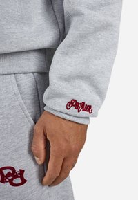 Gray sweatshirt fabric with ribbed cuffs and maroon embroidered logo on the sleeve. Hand rests on matching sweatpants.