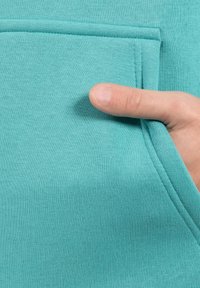 Lyseblå sweatshirt med en tekstureret overflade, der har en forlommekrop. En hånd holder kanten af lommen, hvilket fremhæver dens design.
