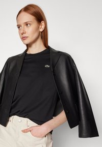 Schwarze Lederblazer über einem schwarzen T-Shirt mit einem kleinen grünen Krokodil-Logo, kombiniert mit hellbeigen Hosen. Glatte Texturen und taillierte Passform.