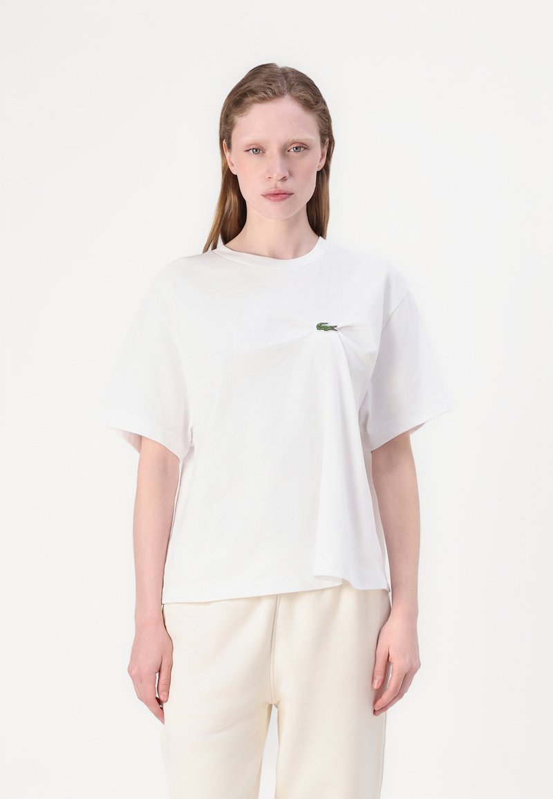 Camiseta blanca de algodón con mangas cortas, de corte holgado, con un pequeño acento de logo verde en el pecho. Textura suave, diseño simple.