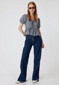 Blusa a maniche corte in gingham bianco e nero con allacciatura frontale, abbinata a pantaloni a zampa d'elefante in denim scuro a vita alta e scarpe di colore chiaro.