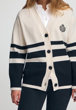 Femme portant un cardigan rayé crème et noir avec quatre boutons et un blason brodé sur une chemise blanche.