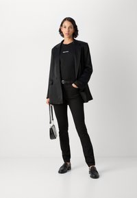 Zwarte op maat gemaakte blazer over een zwart shirt, gecombineerd met zwarte slim-fit broek en zwarte loafers. Houdt een kleine zwarte handtas vast.