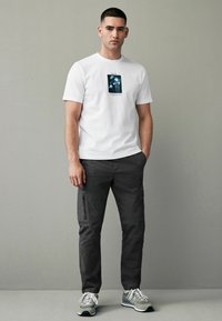 Tricou alb cu imprimeu grafic albastru, asociat cu pantaloni cargo închiși la culoare. Modelul stă în fața unui fundal neutru.