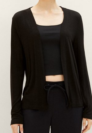 Person iført en sort cardigan med åben front over en sort cropped tanktop og sorte bukser med snøre foran mod en ensfarvet baggrund.