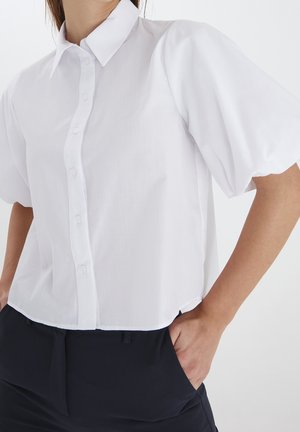 Frau trägt weiße kurzärmelige Bluse mit Puffärmeln und schwarzen Hosen, Hände in den Taschen vor einfarbigem Hintergrund.