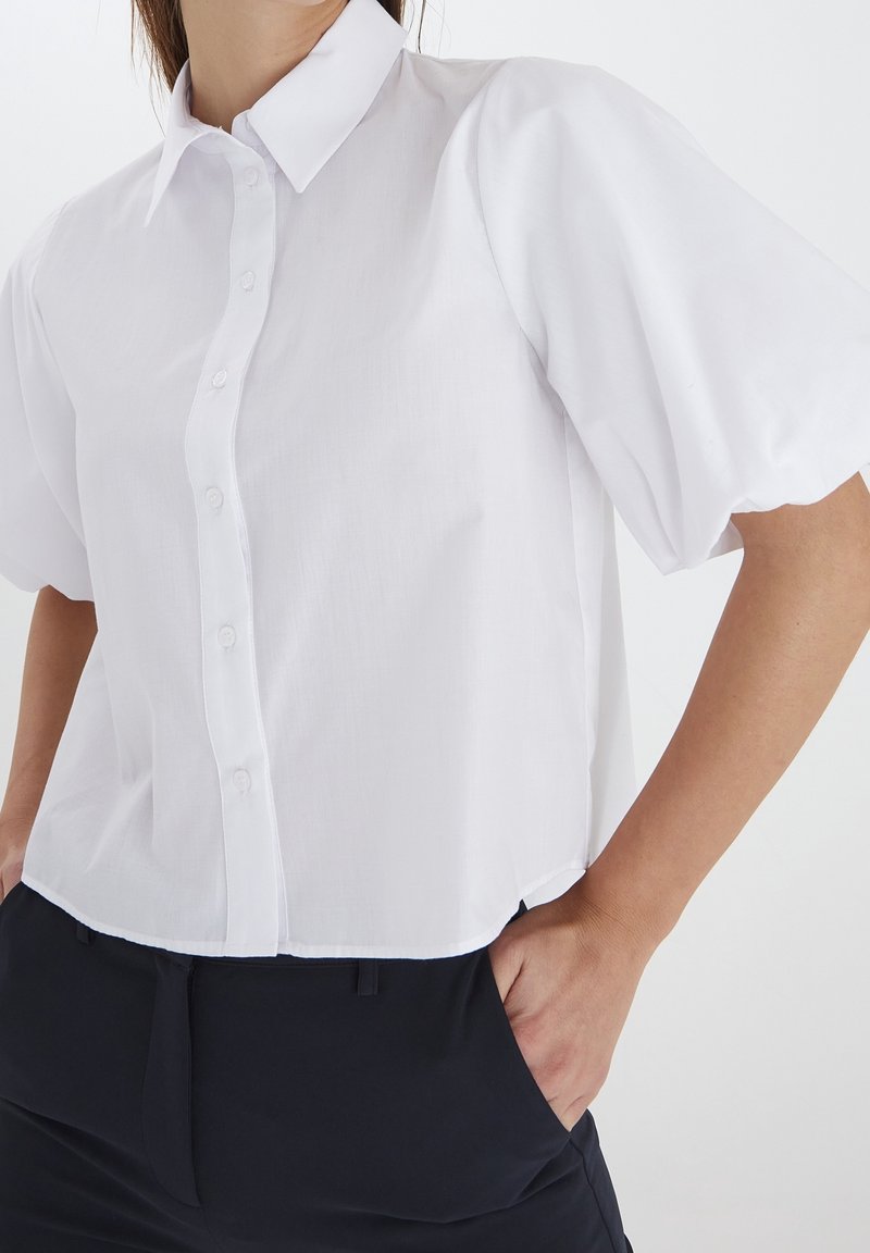 Femme portant un chemisier blanc à manches courtes boutonné avec des manches bouffantes et un pantalon noir, les mains dans les poches, sur un fond uni.
