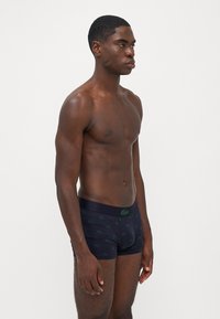 Sötétkék boxeralsók zöld Lacoste logókkal. Puha anyagból készültek, testhezálló szabásúak, elasztikus derékkal rendelkeznek, és nincsenek jól látható mintáik.