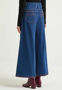 Pantaloni in denim a gamba larga di colore blu scuro. Presentano cuciture arancioni a contrasto e due tasche posteriori con disegni a zigzag unici.