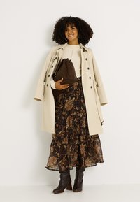 Trench-coat beige, pull à côtes crème, jupe maxi fleurie dans des tons sombres, sac marron décontracté et bottes à talons noires.