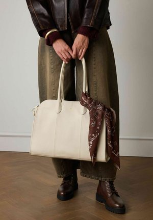 Sac en cuir crème avec deux poignées et une écharpe en paisley marron nouée. Modèle portant un pantalon ample vert olive et des bottes en cuir lacées marron foncé.