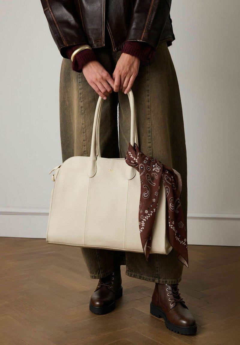 Crème leren handtas met twee handvaten en een bruine paisley sjaal eromheen geknoopt. Model draagt losse olijfgroene broek en donkerbruine veterschoenen.