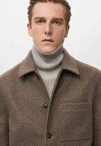 Brauner Mantel aus Wollmischung mit Hemdkragen, einer einzelnen Brusttasche und schwarzen Knöpfen; darüber getragen ein hellgrauer, gerippter Rollkragenpullover.