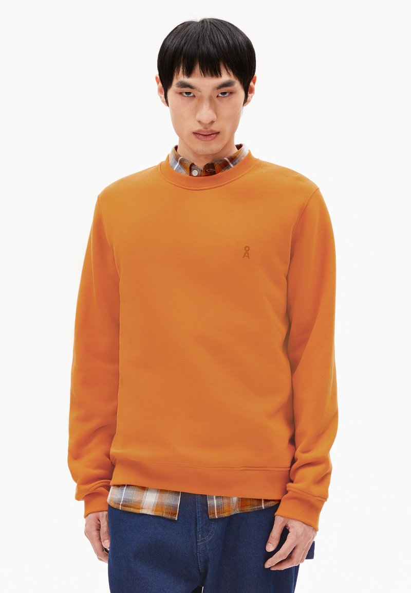 Felpa arancione con scollo rotondo, maniche lunghe e un piccolo logo. Indossata sopra una camicia a quadri e abbinata a jeans in denim.
