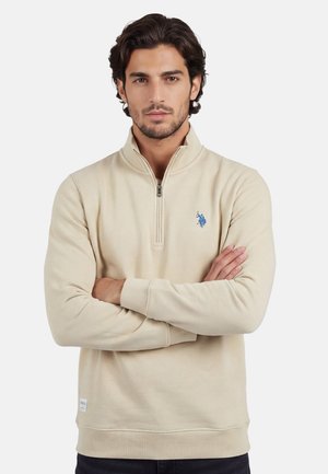 Mann mit dunklen Haaren, der einen beigen Pullover mit Viertel-Reißverschluss und blauem Logo trägt, die Arme verschränkt, blickt nach vorne vor einem weißen Hintergrund.