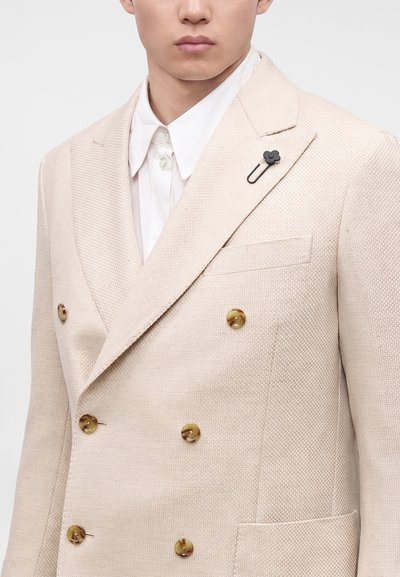 Homme portant un blazer beige texturé à double boutonnage avec des boutons en écaille de tortue et une chemise blanche à col en dessous.