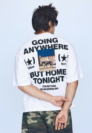 Homme aux cheveux foncés portant des lunettes de soleil, un t-shirt blanc avec un motif graphique, du texte et une photo de cheval, et un pantalon camouflage, vu de dos.
