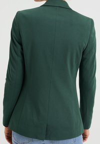 Grön skräddarsydd kavaj med en slim fit, flikiga lapeller och en enda ventil bak. Slät tyg med matt yta, minimal sömnadsdetalj.