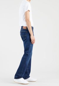 Personne debout de profil, portant un jean bleu, un t-shirt blanc et des baskets blanches sur un fond clair uni.