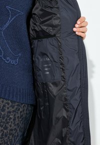 Osoba držící otevřenou černou bundu s viditelnou vnitřní kapsou označenou jako voděodolná, větruodolná a prodyšná tkanina.