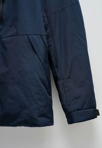 Veste isolante bleu marine, avec un design élégant, un tissu texturé et un poignet ajustable en velcro pour un meilleur ajustement. Poche latérale visible sur la manche.