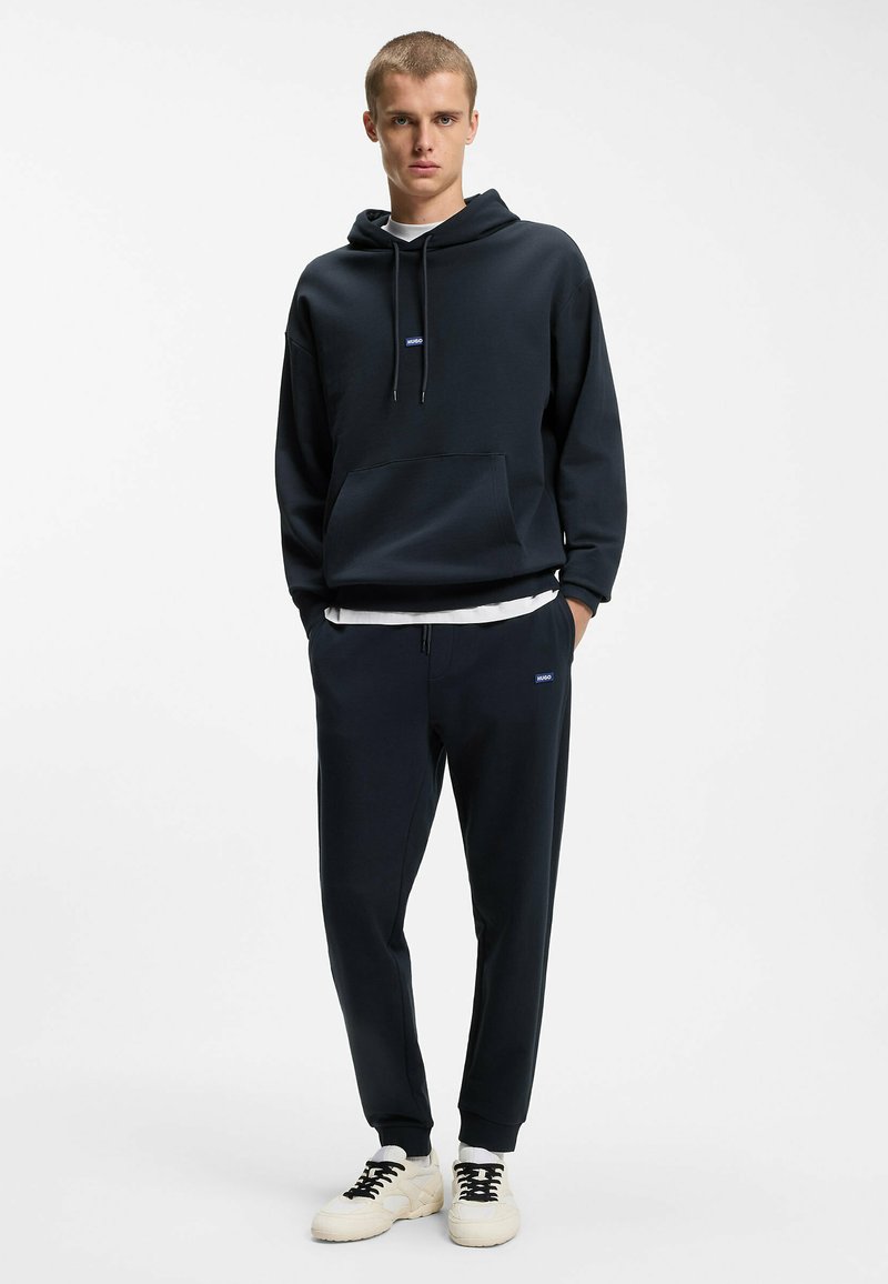 Jeune homme debout portant un sweat à capuche noir et un pantalon de jogging avec des baskets blanches sur un fond blanc uni.