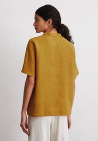 Mustard yellow linen short-sleeve shirt with a straight cut and a back seam. Worn with light-colored trousers. Minimalist design.