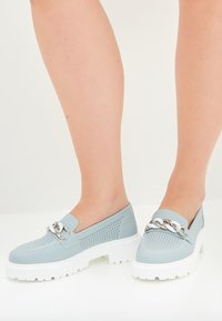 Světle modré slip-on boty s perforovaným designem, robustní bílou podrážkou a stříbrným řetízkem na nártu. Hladká textura a zaoblený tvar.