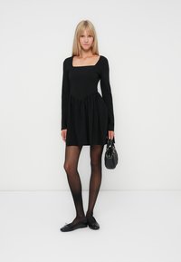 Vestido negro de manga larga con escote cuadrado y falda fruncida. Llevado con medias negras transparentes y zapatos negros planos. Pequeño bolso como acento.