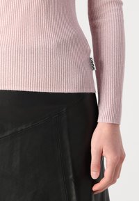 Pull en maille rose clair à manches longues, avec un éclat métallique et une petite étiquette noire "HUGO" sur le côté, associé à une jupe en cuir noir.